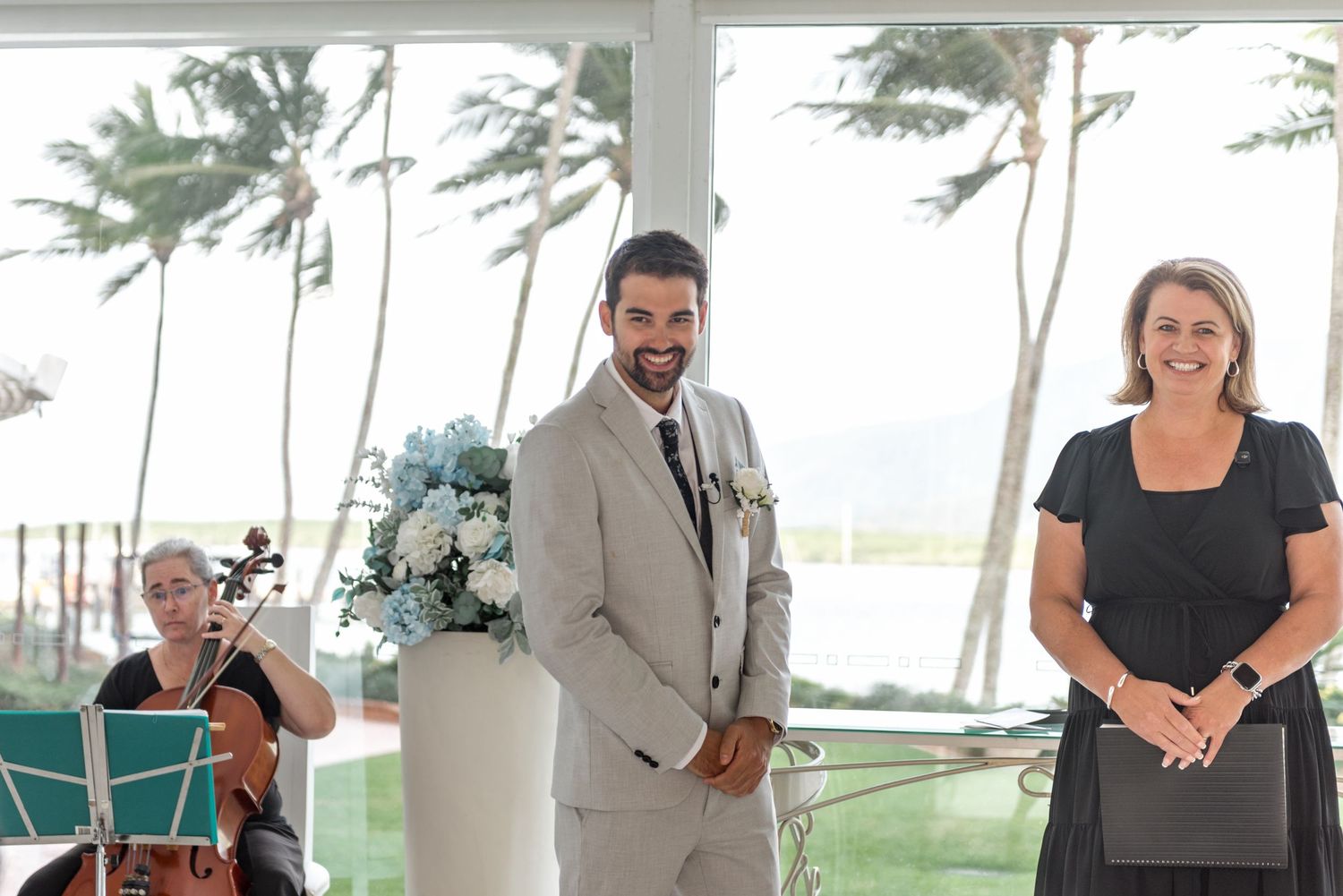 Smiling groom in grey suit stands at altar with celebrant and cellist at tropical wedding.