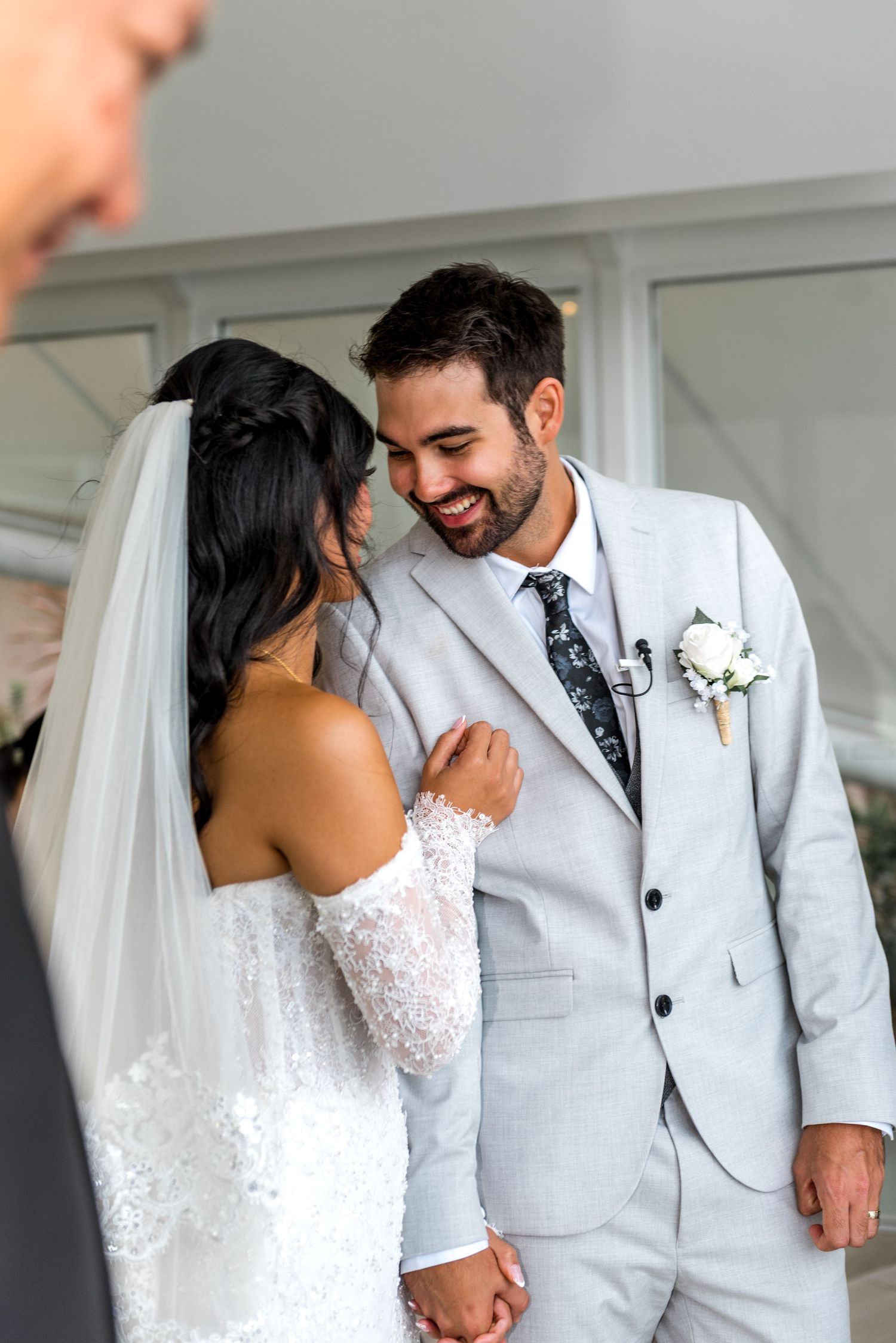 Smiling groom in light gray suit holds bride's hand during intimate wedding ceremony moment.