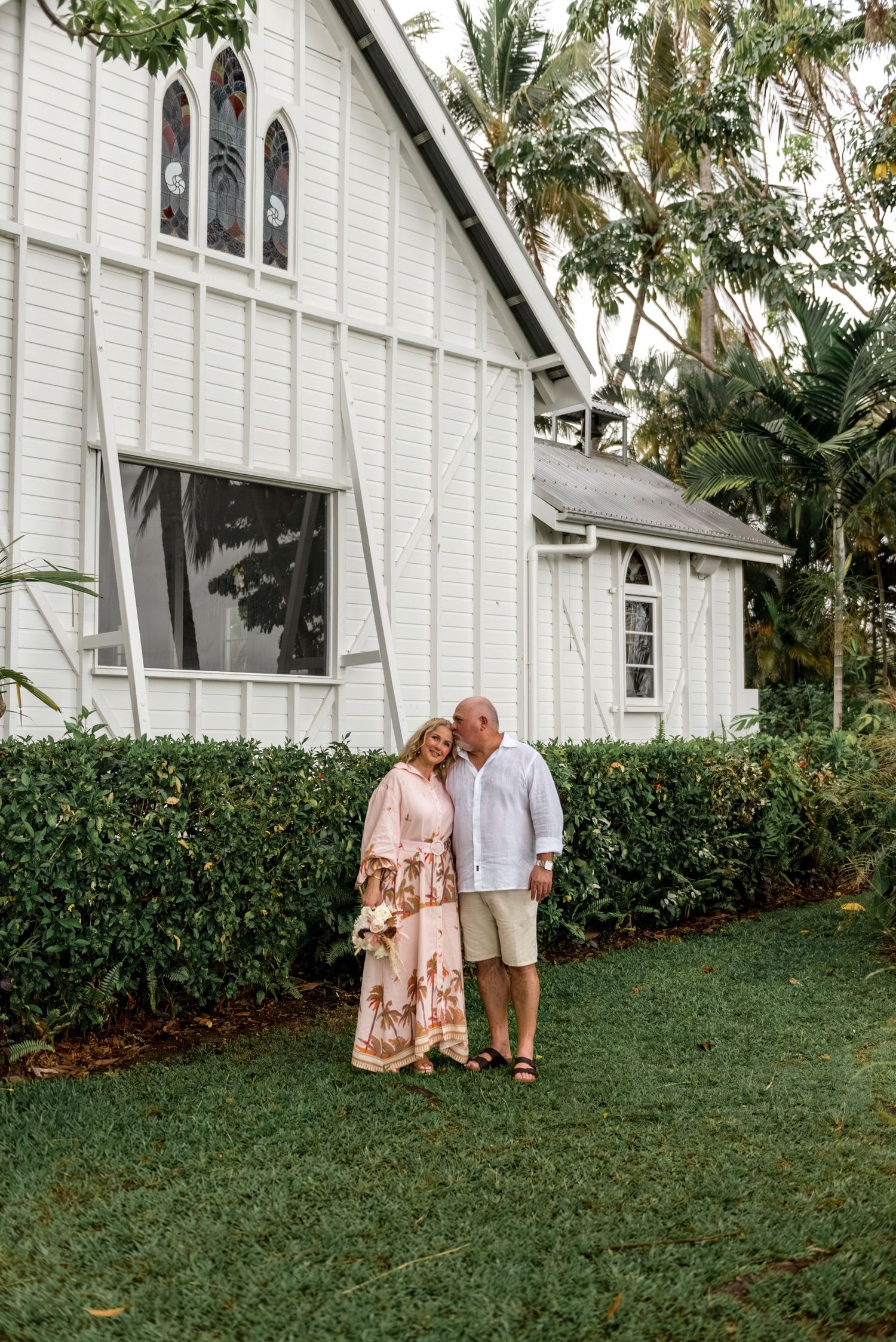 Couple posing affectionately in front of a historic white timber chapel surrounded by greenery.