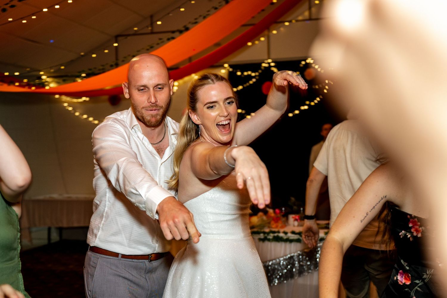 Excited bride in white dress dances energetically with groom at a lively wedding reception.