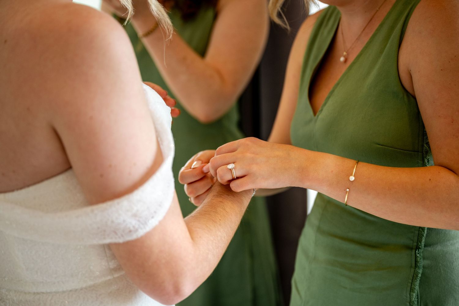 Bridesmaid in green dress helping bride with ring during wedding preparations.