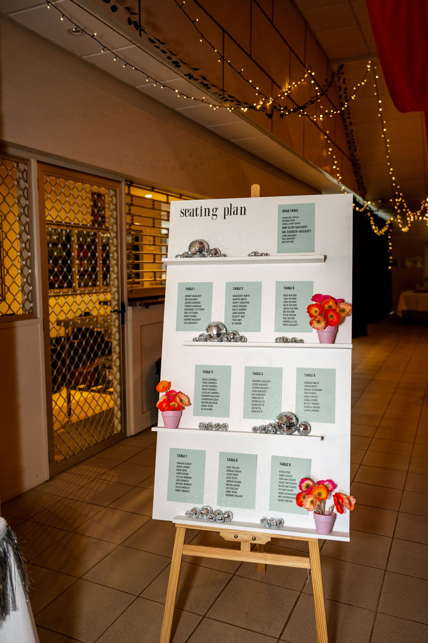 Wedding seating plan displayed on white board on wooden easel decorated with small floral arrangements and disco balls.