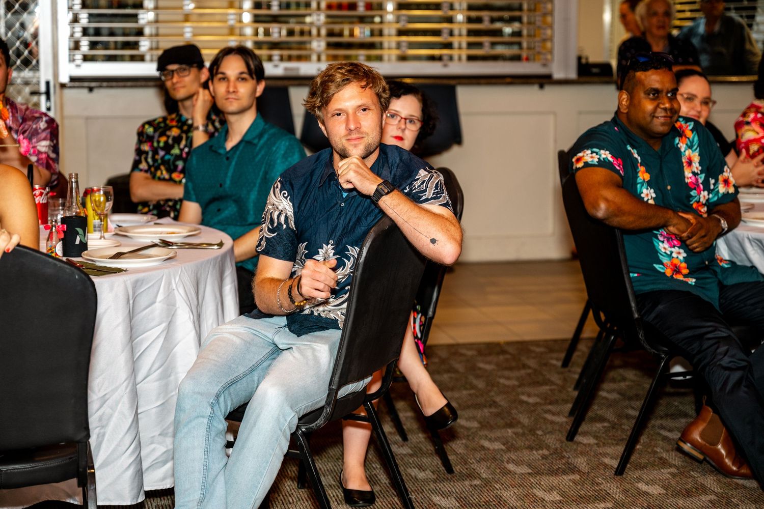 Guests seated at round tables during a wedding reception, attentively watching a presentation in a decorated venue.