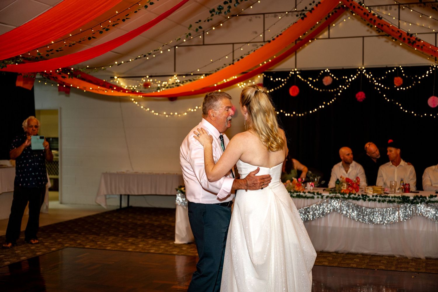 Bride and groom share their first dance at a decorated wedding reception with fairy lights and red draping.