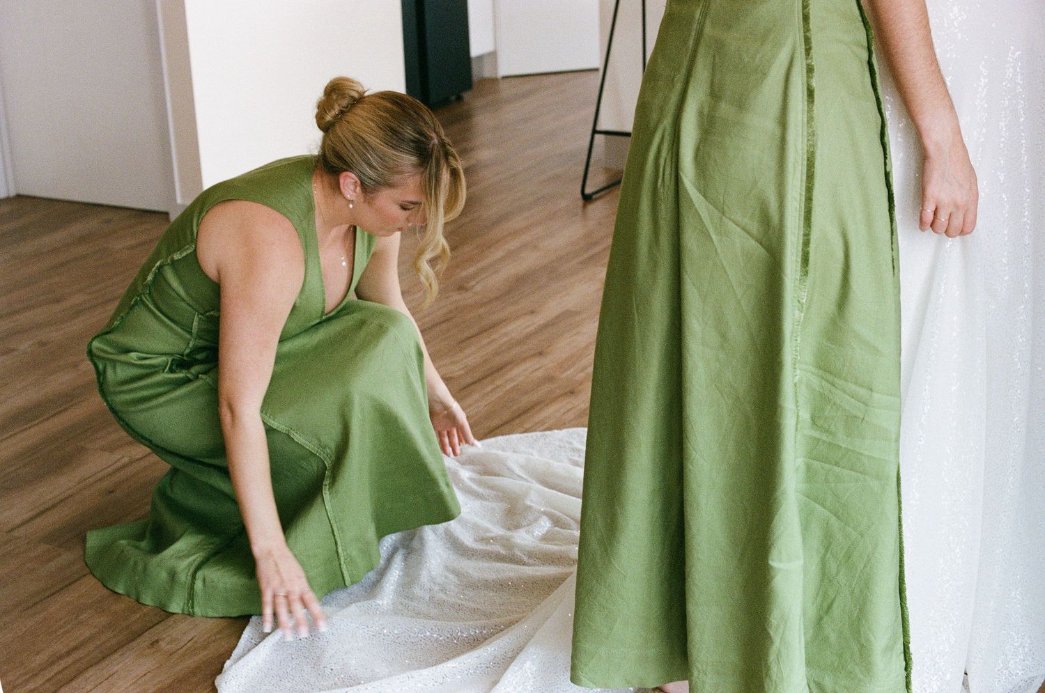 Bridesmaid in green satin dress adjusts bride's white wedding gown train on wooden floor.