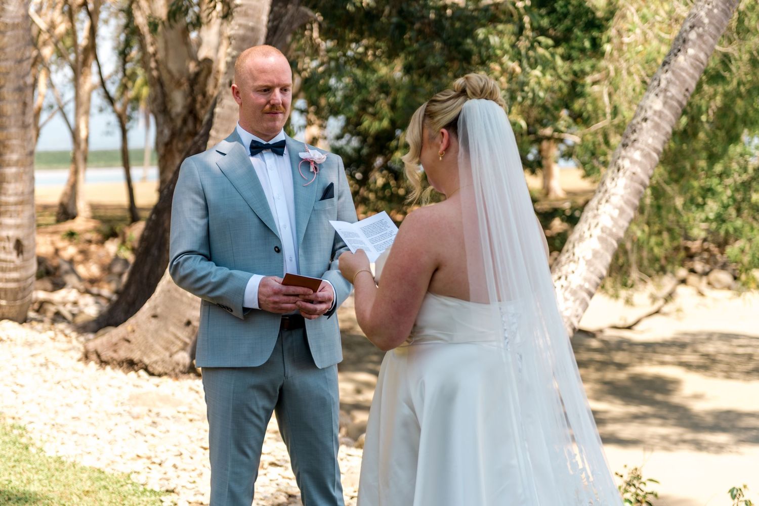 Groom reads vows holding book at tropical outdoor wedding ceremony with bride in white gown.