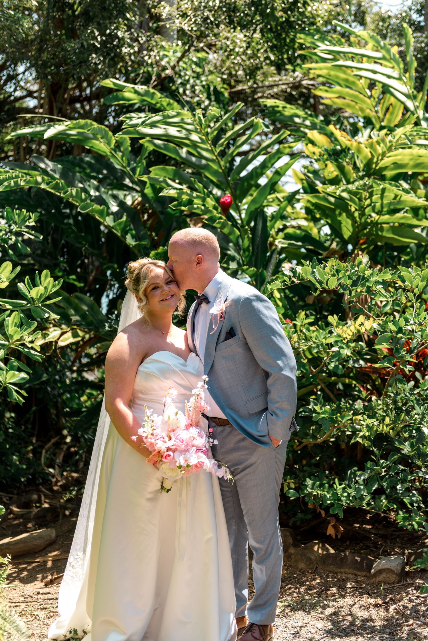 Bride and groom smile together in front of lush tropical garden greenery, bride holding a pink bouquet.