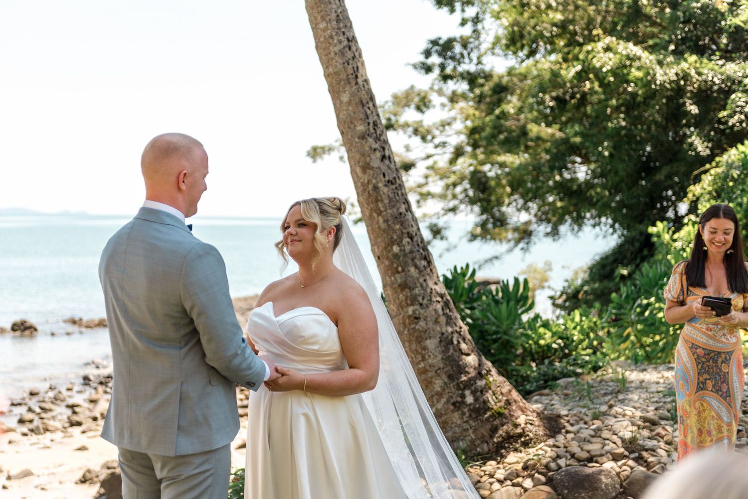 Bride and groom exchange vows at beachside ceremony with officiant holding book beside palm tree.