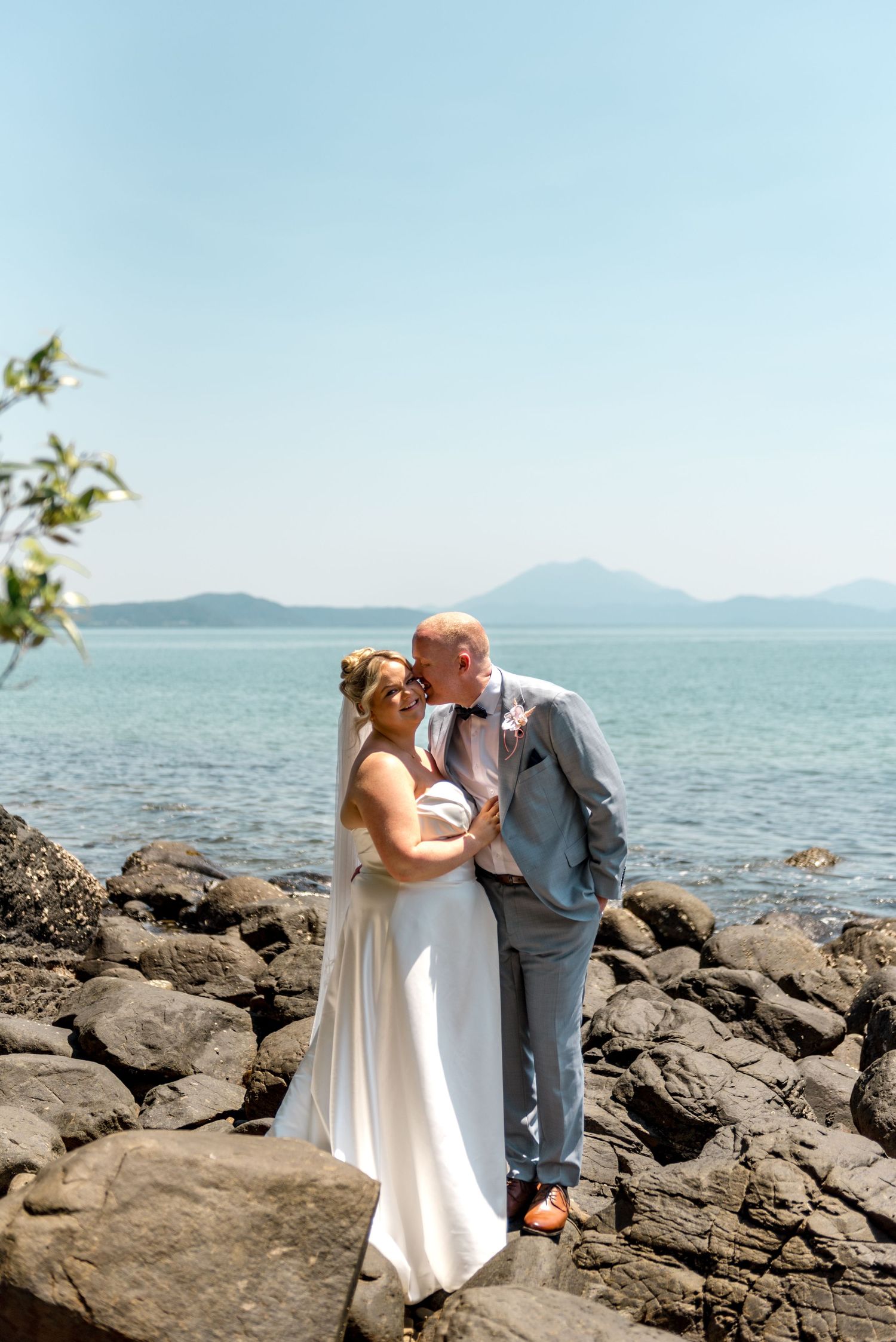 Groom kisses bride on rocky oceanside shore with blue water and mountain silhouettes in background.