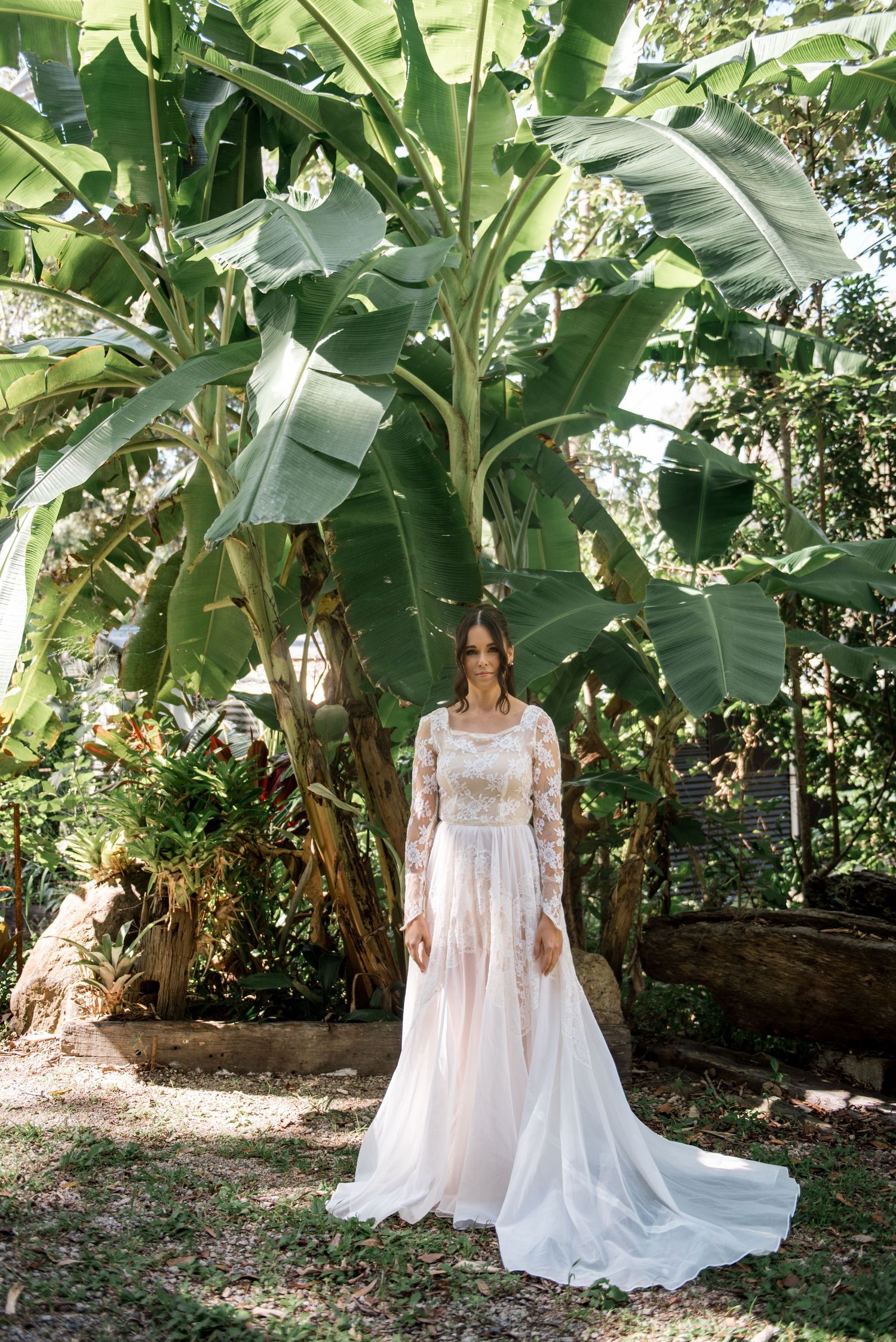 Bride in lace long-sleeve wedding gown standing beneath tropical banana tree outdoors.