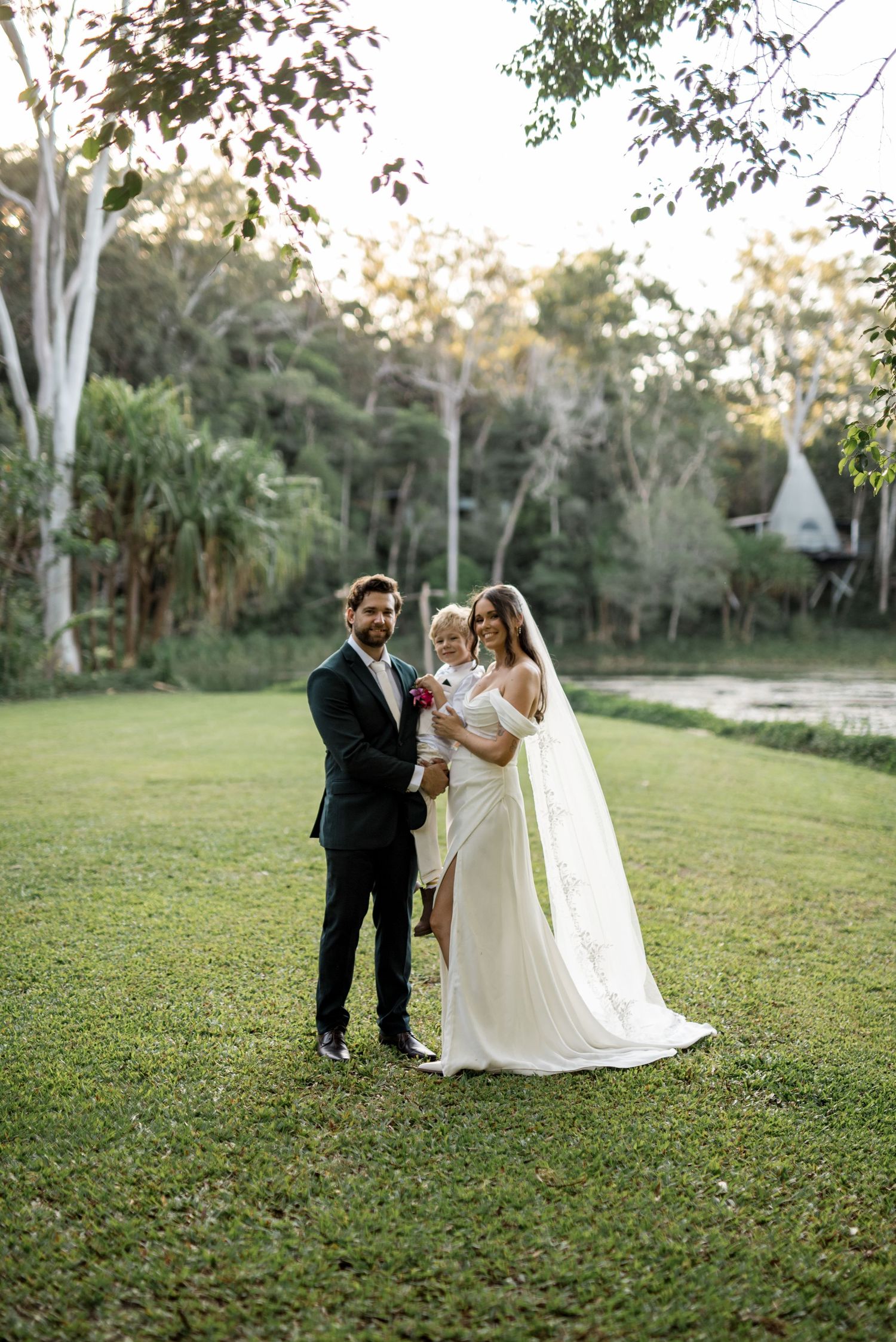 Bride and groom in wedding attire hold a young child on a lush green lawn outdoors.