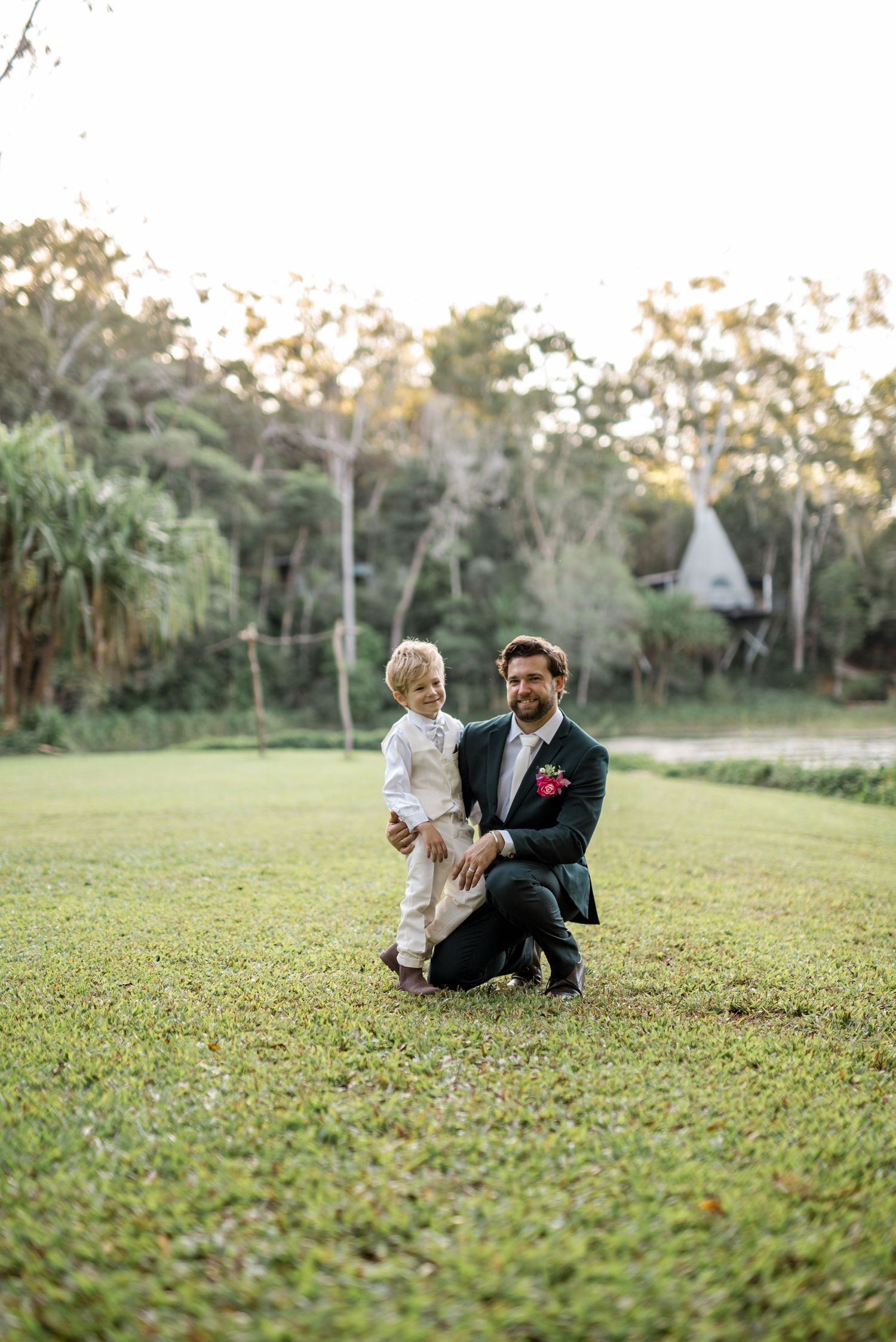 Groom in dark suit kneels beside a young blond boy on a green lawn with trees behind.