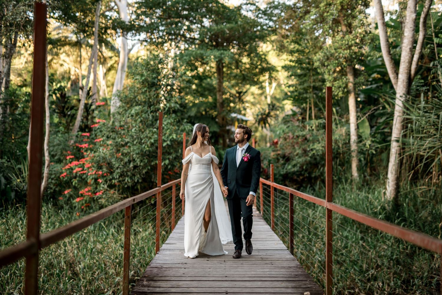 Bride and groom walk hand-in-hand across a wooden bridge through a lush tropical garden.