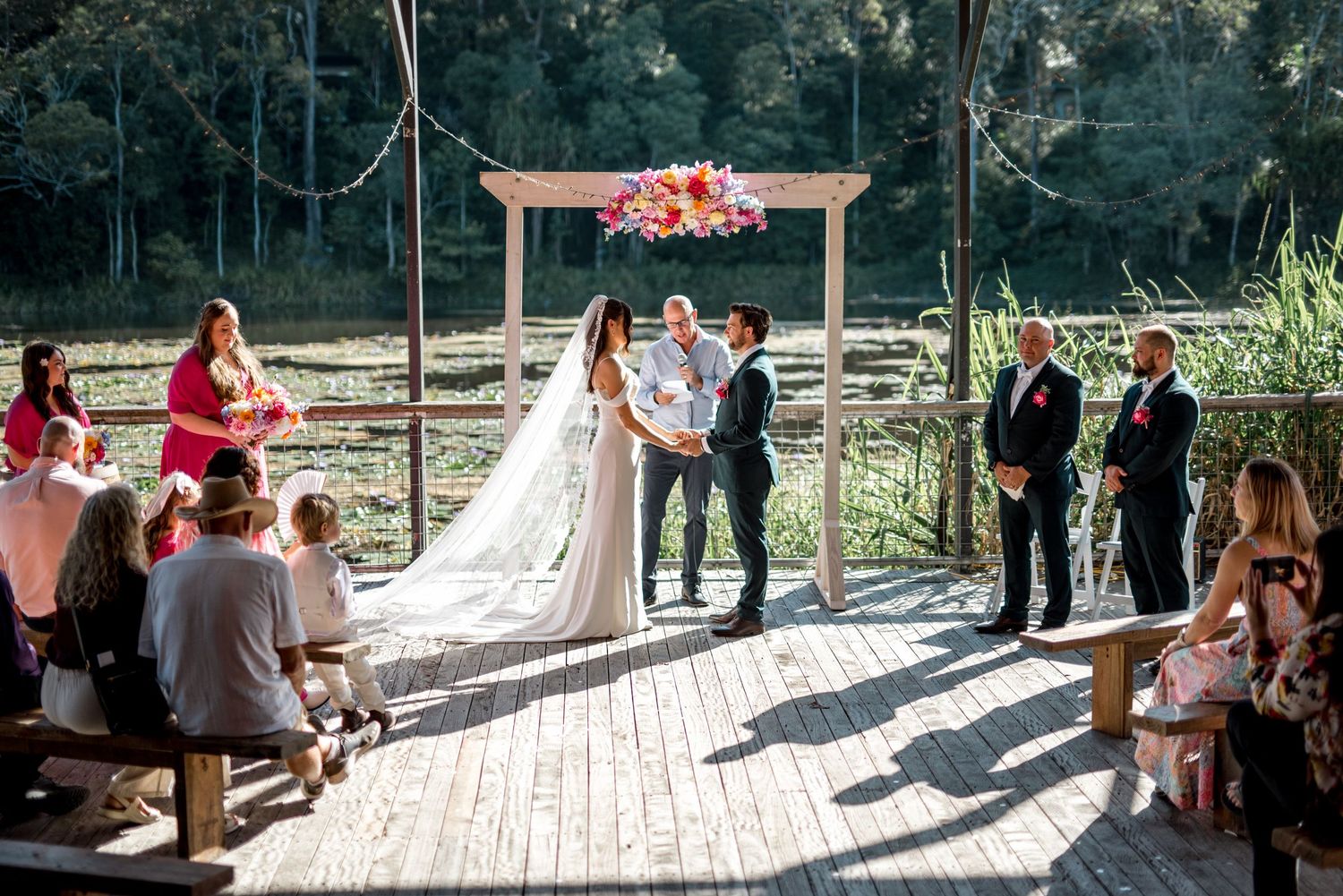 Outdoor wedding ceremony on wooden deck with bride and groom at floral arch, guests seated on rustic benches by a river.