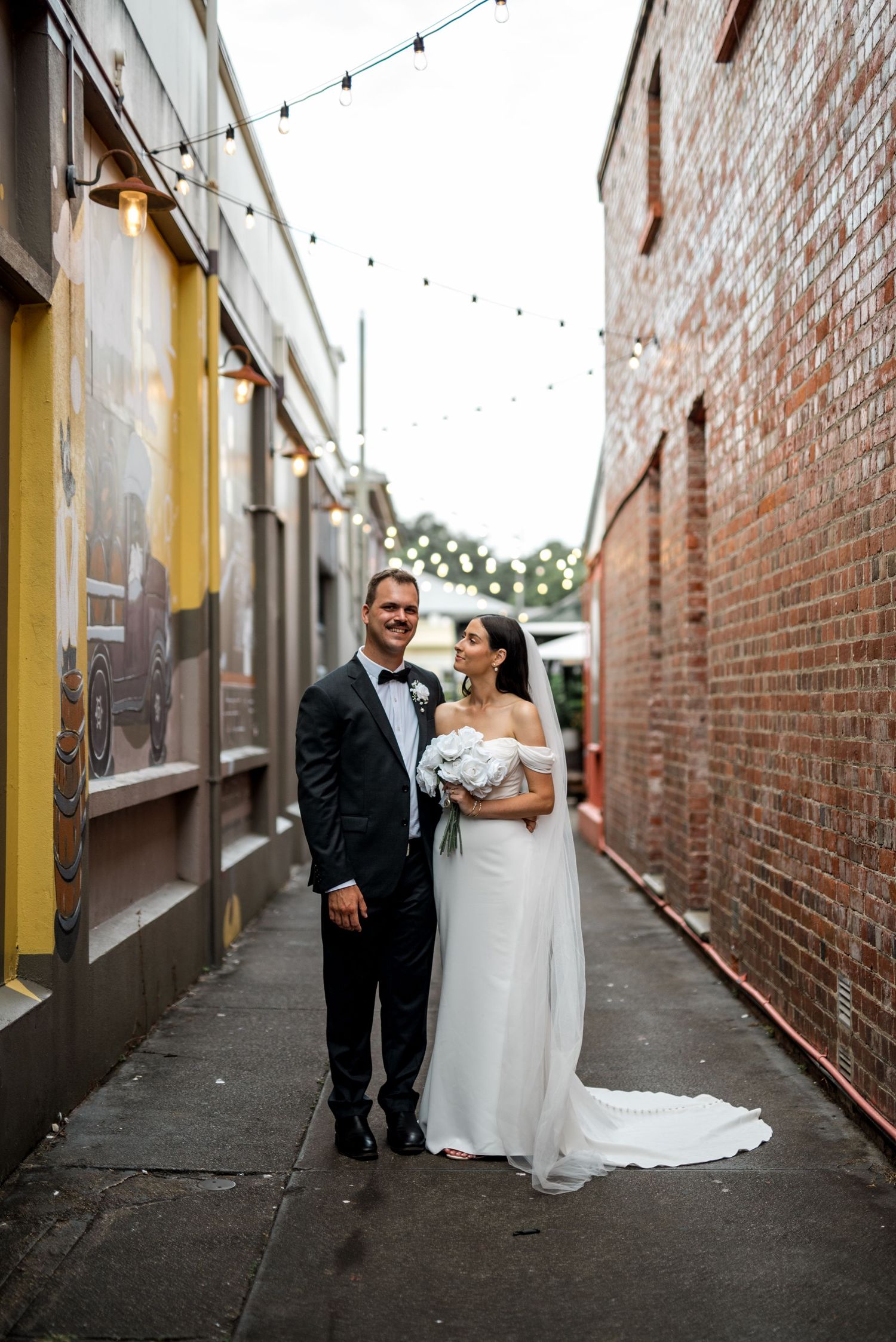 Bride and groom pose in a charming brick alley adorned with string lights on their wedding day.