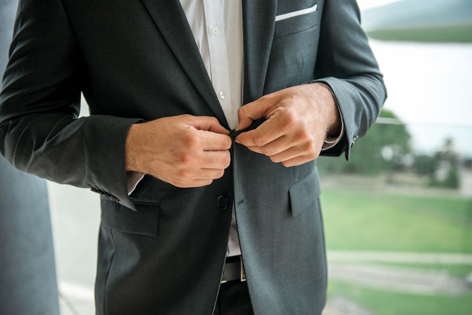 Close-up of groom buttoning his dark suit jacket with a waterfront view visible through the window behind him.