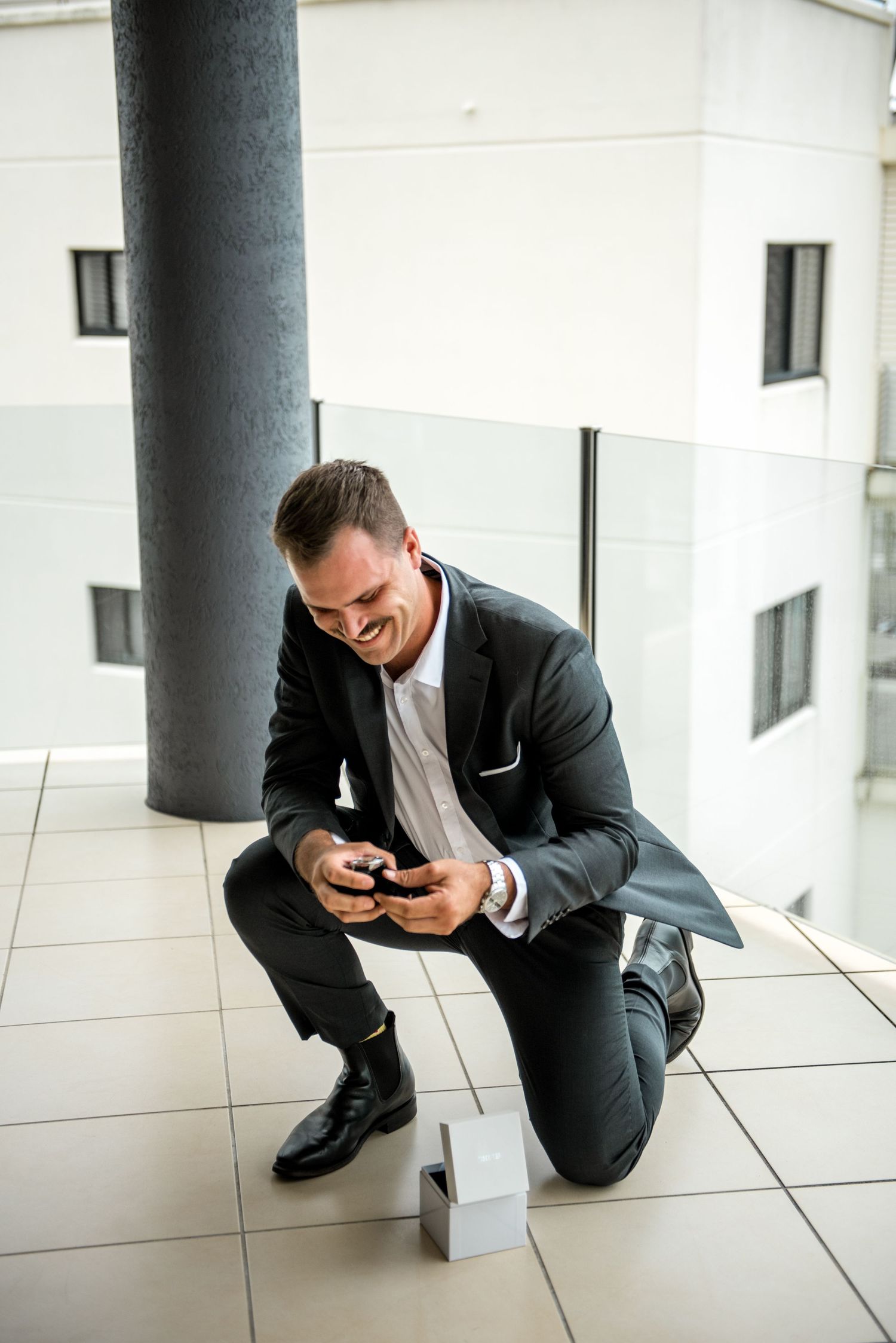 Smiling groom in dark suit kneels on balcony floor reading a message on his phone before wedding.
