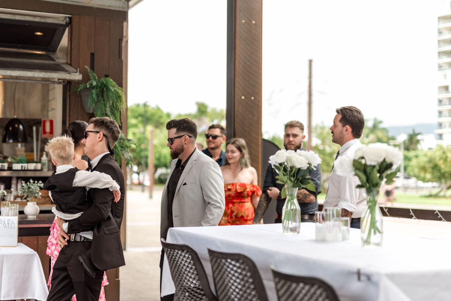 Wedding guests mingling indoors at a reception venue with white rose centerpieces on draped tables.