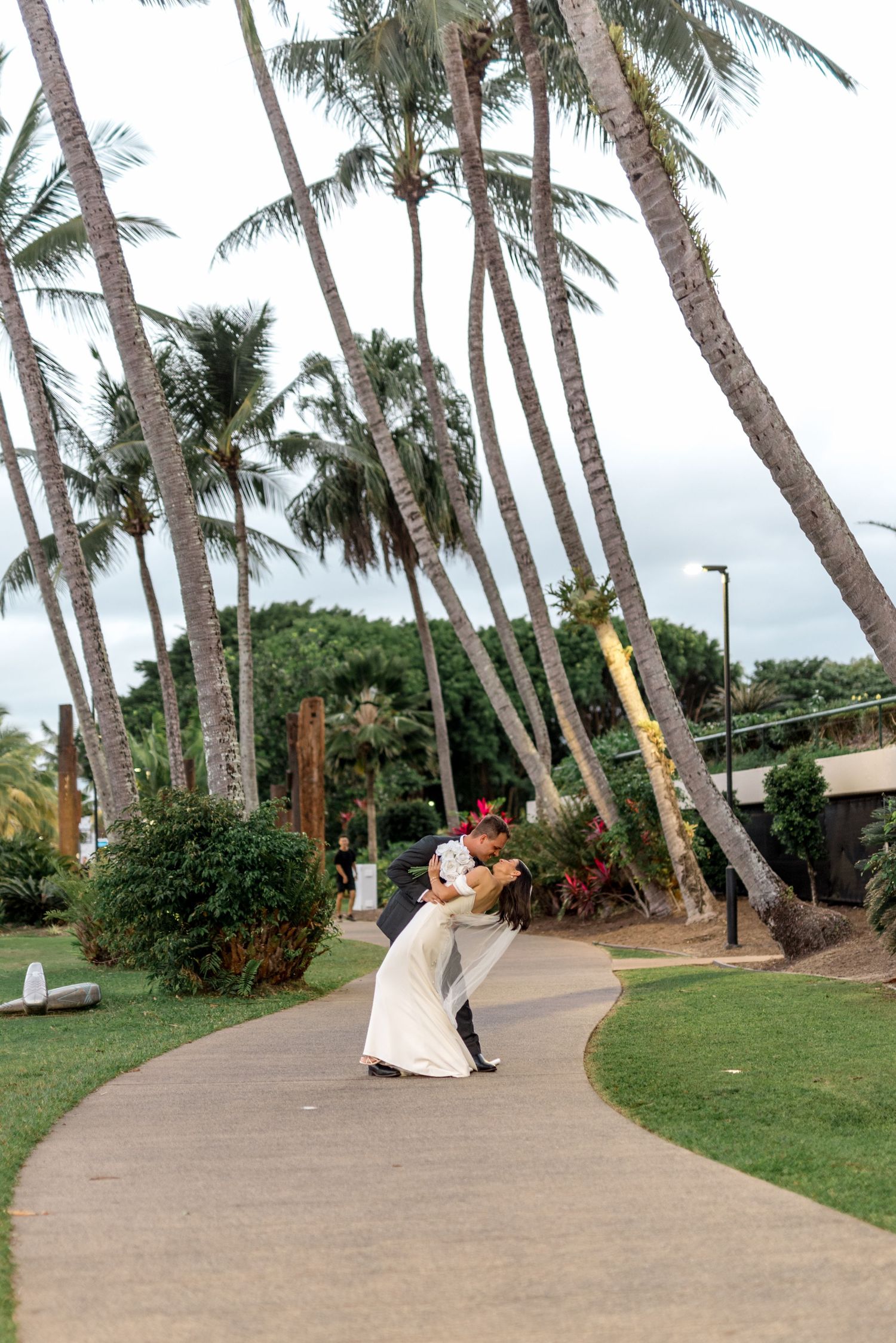 Groom dipping bride in a romantic pose on a curved garden path lined with tall swaying palm trees in Hawaii.