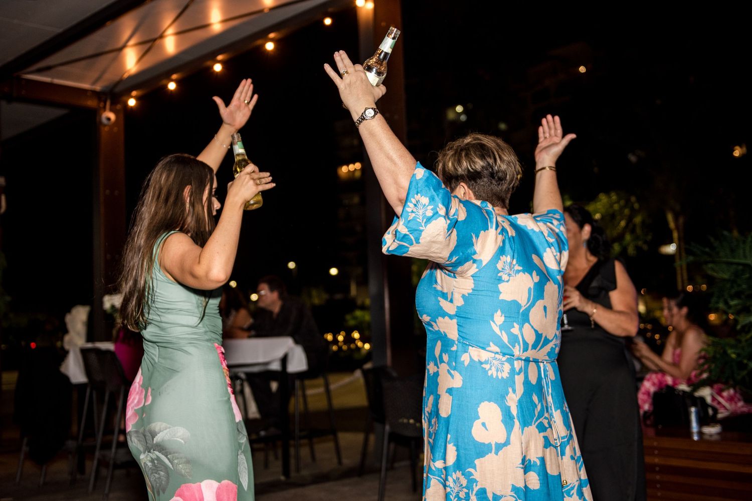 Two women raise their hands joyfully while dancing at an outdoor evening wedding reception.