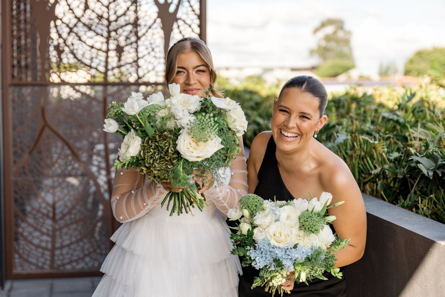 Bride and bridesmaid in black dress smile holding white and blue floral bouquets at venue.