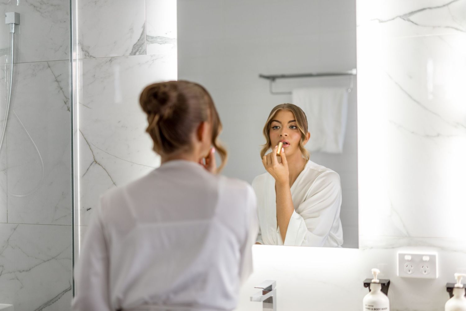 Bride in white robe applying lipstick while looking in illuminated bathroom mirror during wedding prep.