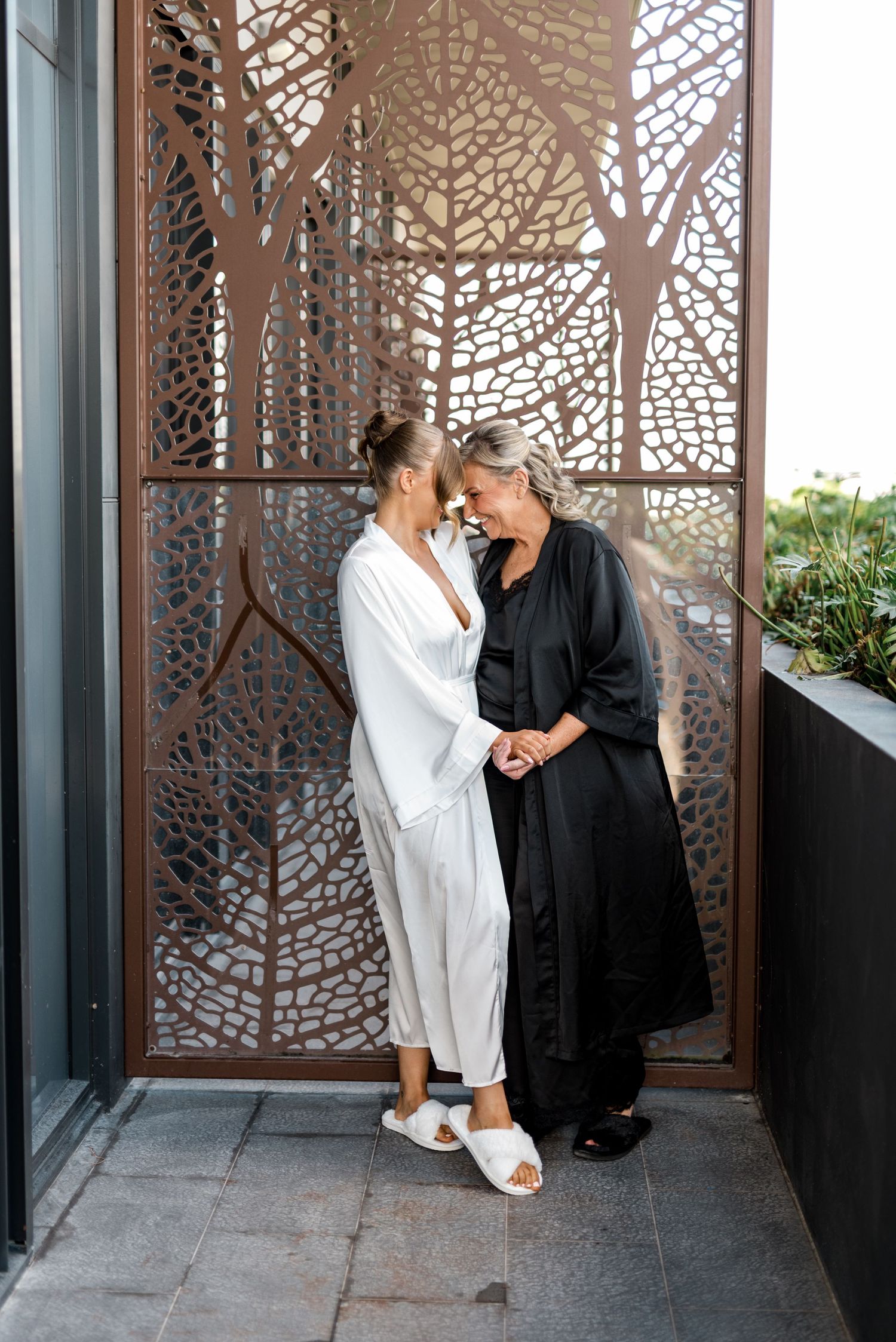 Bride in white satin robe sharing an intimate moment with older woman in black robe on hotel balcony.