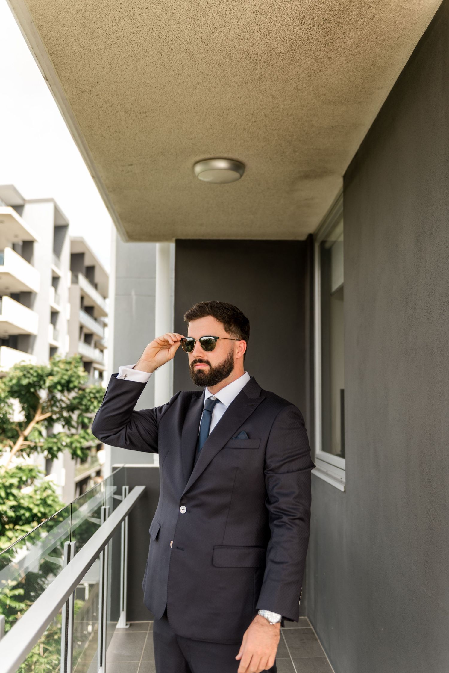 Confident groom in a navy suit and sunglasses posing on a balcony on his wedding day.