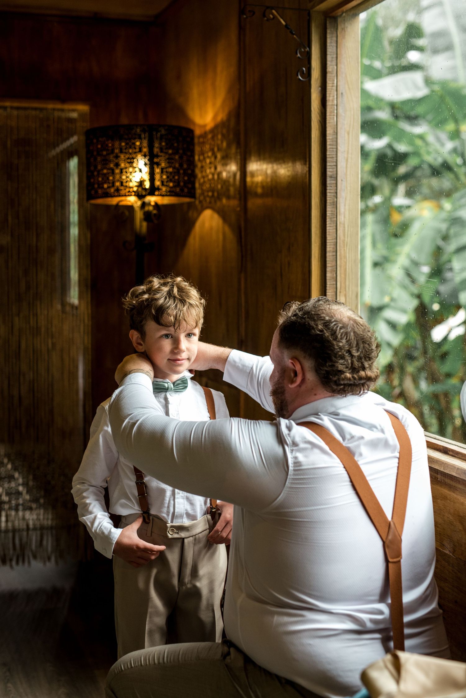 Groom kneeling to adjust green bow tie on young ring bearer in beige suit by a sunlit window.