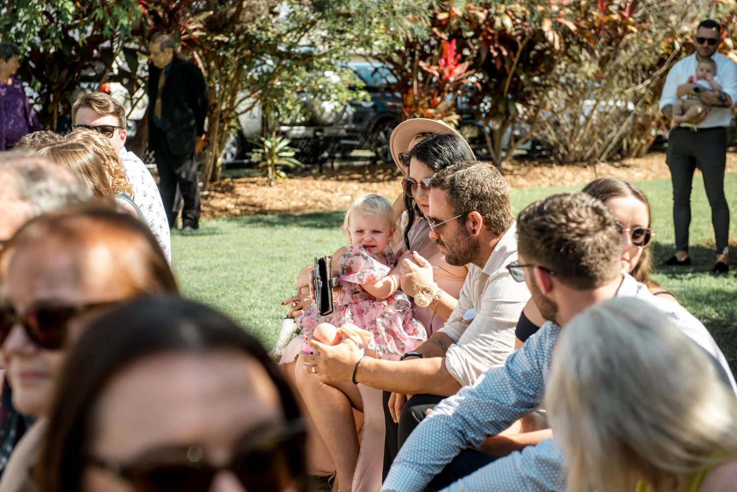 Wedding guests seated outdoors on grass, man holding a baby girl in a floral dress in sunlight.
