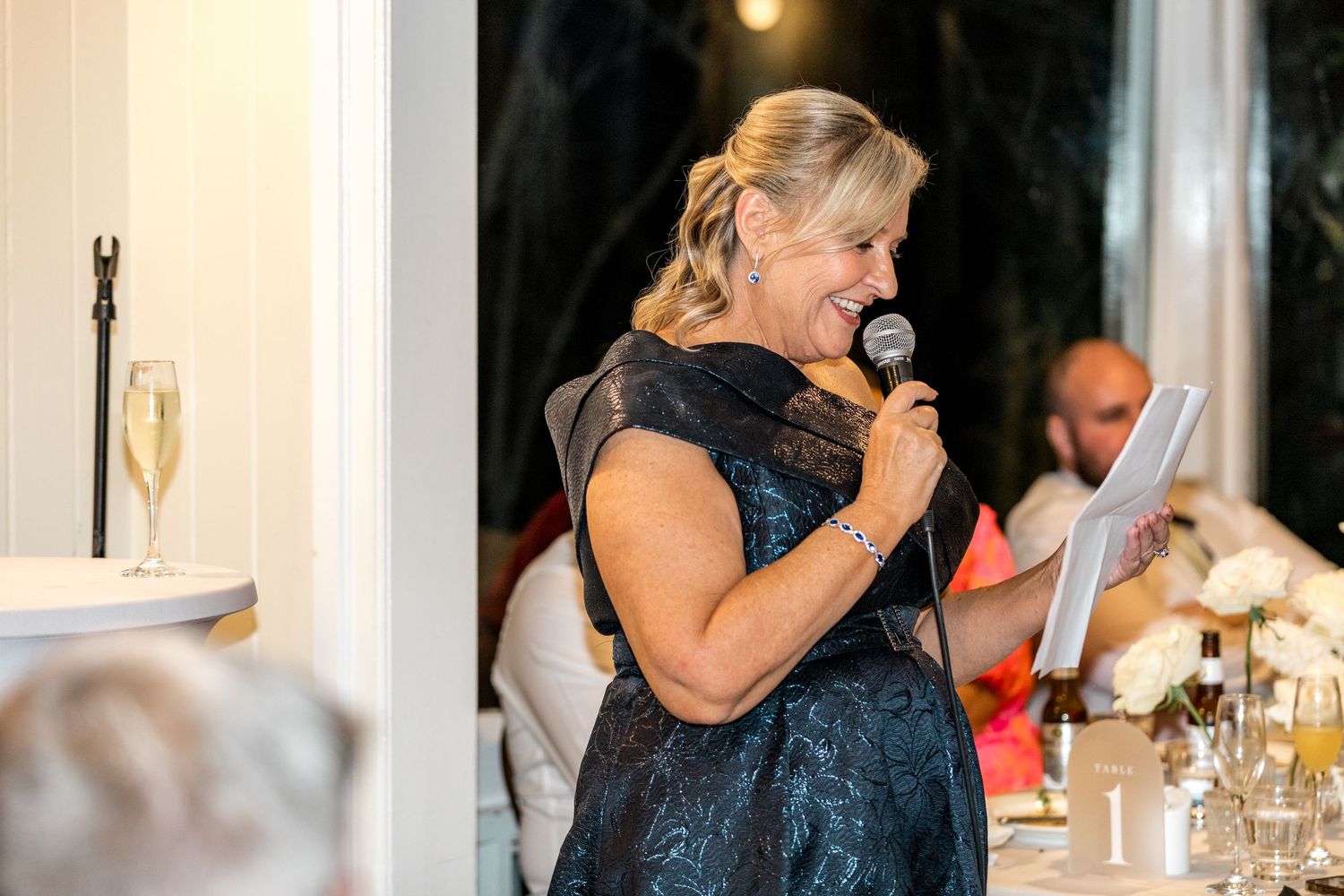 Woman in blue lace dress giving a heartfelt speech at an elegant wedding reception.