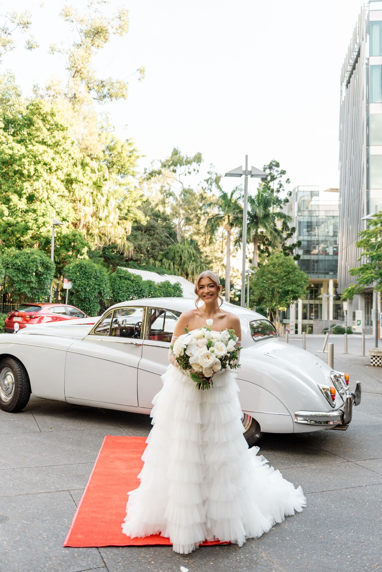 Bride in ruffled white gown holds bouquet beside a vintage white classic car.