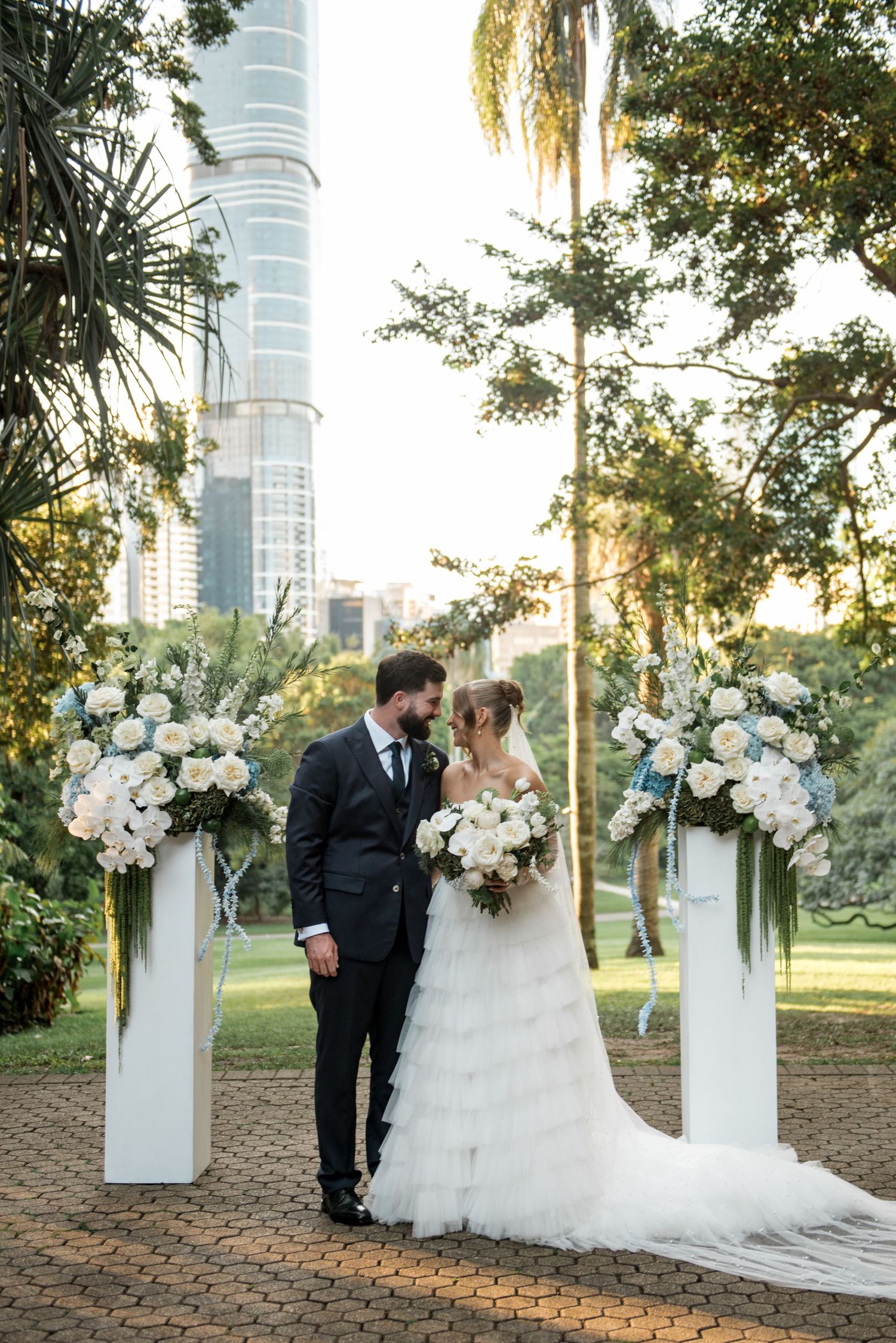 Newlyweds embrace at elegant outdoor wedding ceremony with white rose arrangements and city backdrop.