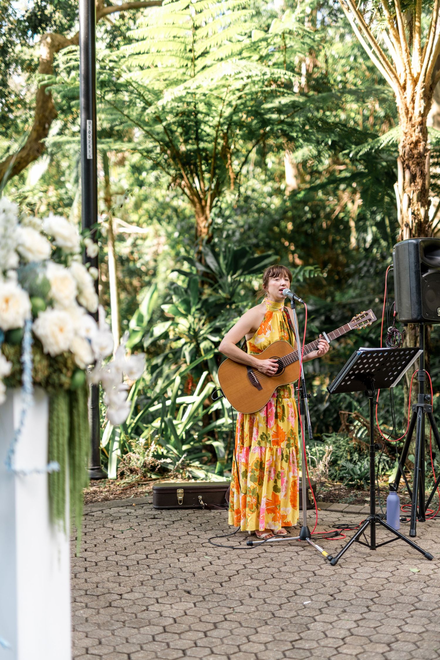 Female musician in floral maxi dress playing acoustic guitar at an outdoor garden wedding ceremony.