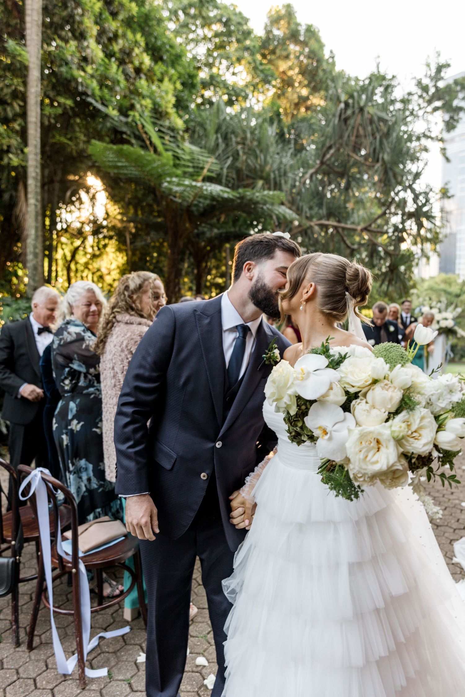 Newlyweds share a kiss while walking down the aisle, bride holding a white peony bouquet.