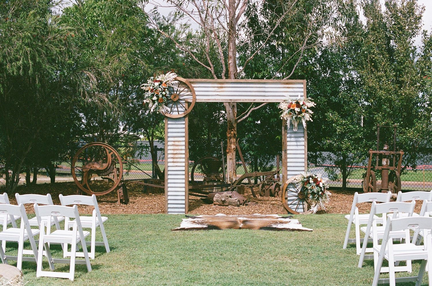 Outdoor rustic wedding ceremony setup with white chairs, wooden arch, and wagon wheel decorations.