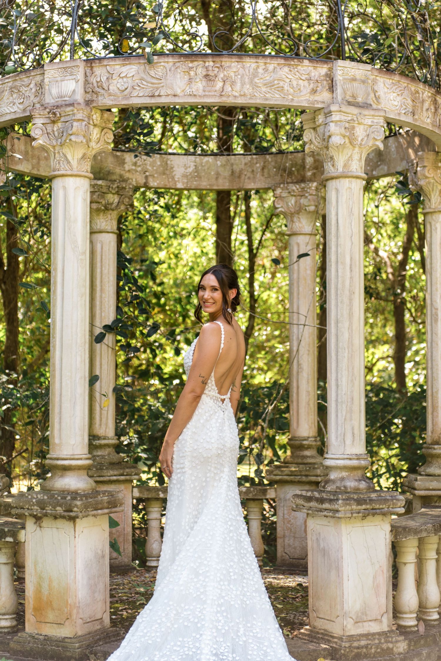 Bride in low-back sequined gown with cathedral train poses gracefully inside stone columned garden gazebo.