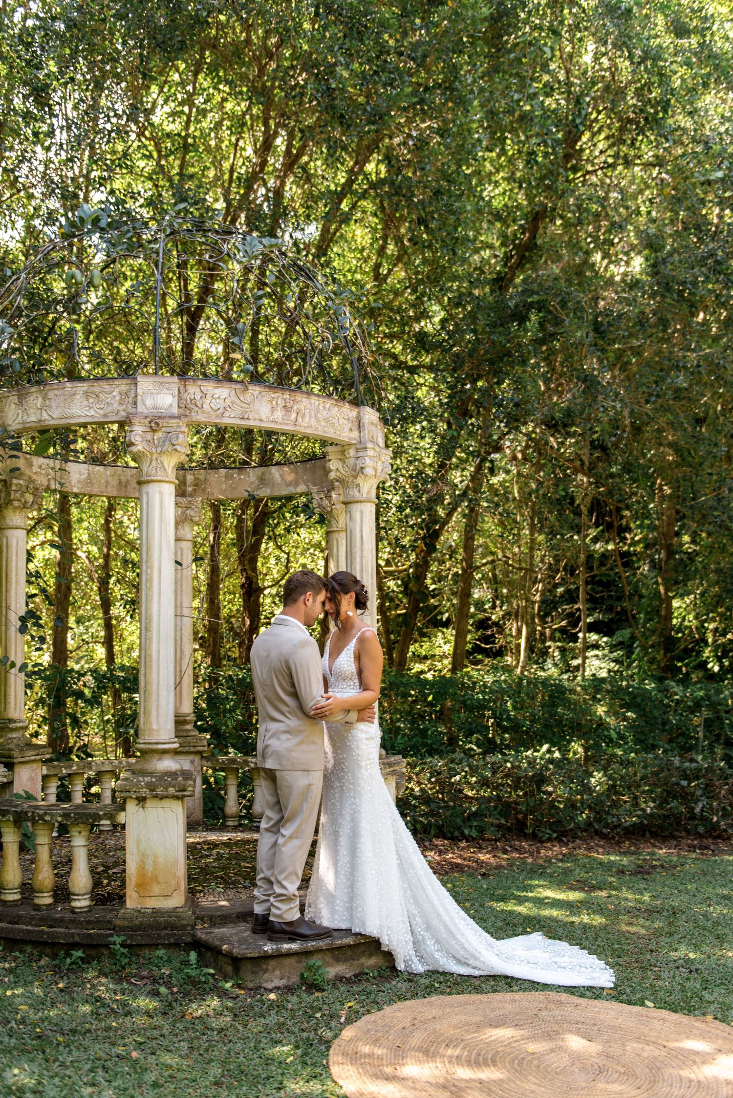 Bride and groom sharing intimate moment under ornate stone gazebo surrounded by lush green garden trees.
