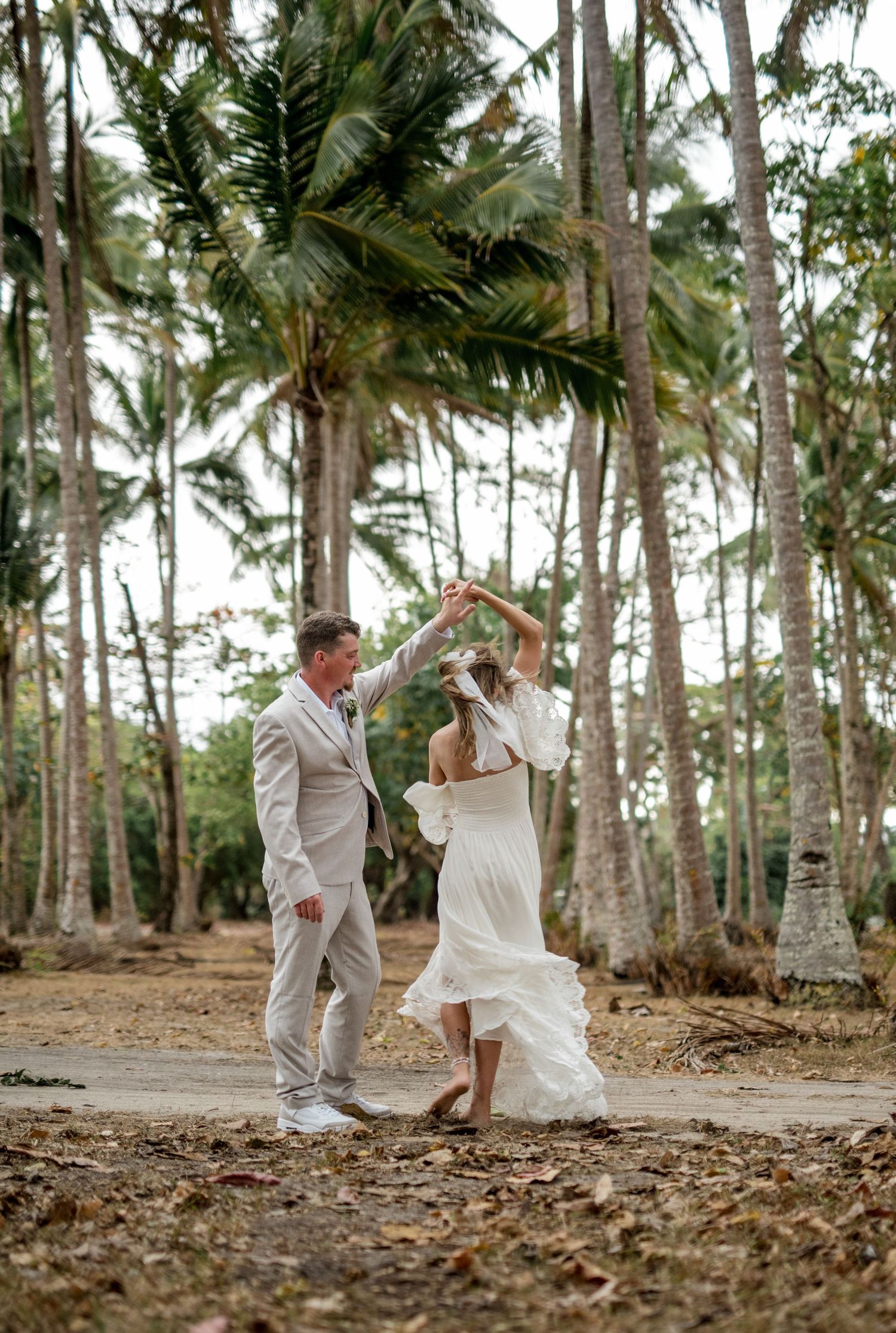 Groom spins bride in flowing white dress under tall coconut palms during tropical wedding celebration.