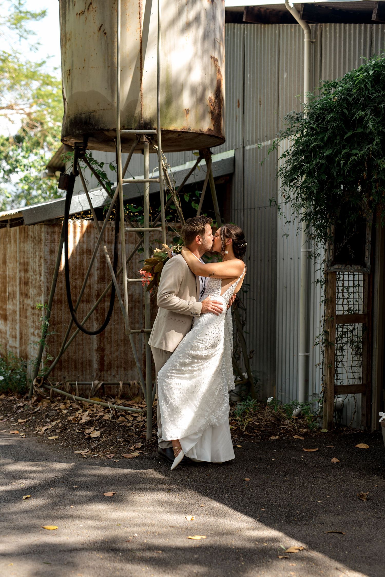 Bride and groom share a passionate kiss beside a rustic industrial structure outdoors.