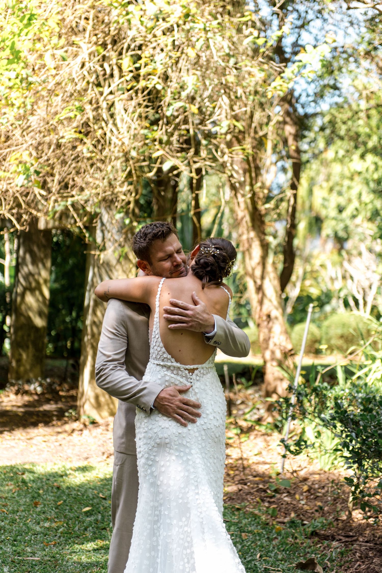 Bride and groom sharing tender embrace in sunlit garden, showcasing bride's elegant open-back wedding gown.