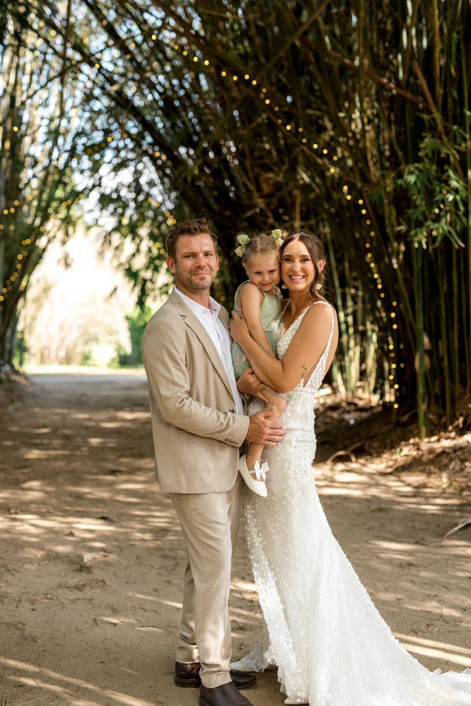 Smiling bride and groom holding their young daughter under a bamboo tunnel with fairy lights.