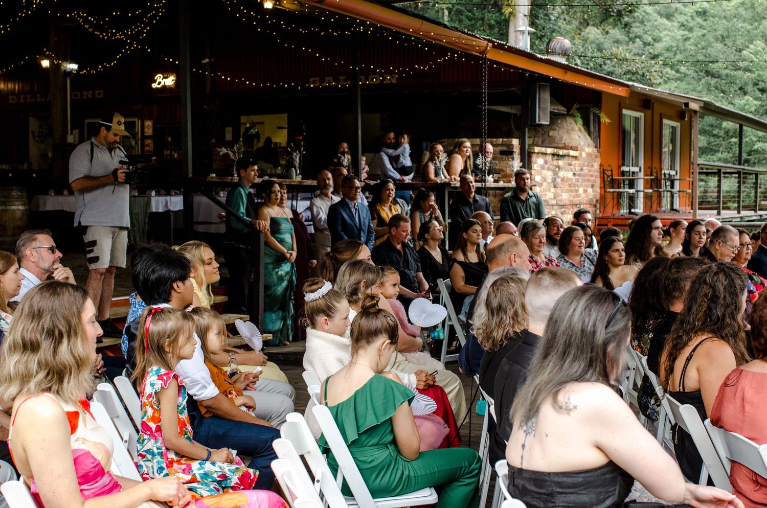 Large wedding reception crowd seated outdoors under string lights at rustic venue.
