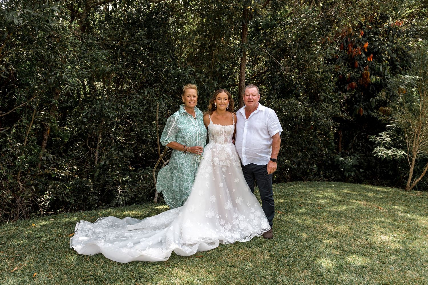 Bride in floral lace gown poses with two guests in a lush green garden on her wedding day.