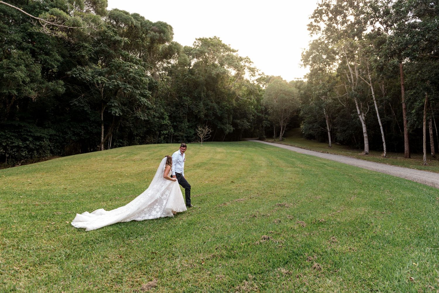 Bride in flowing white gown and groom share a tender moment on a green lawn beside a forest path.