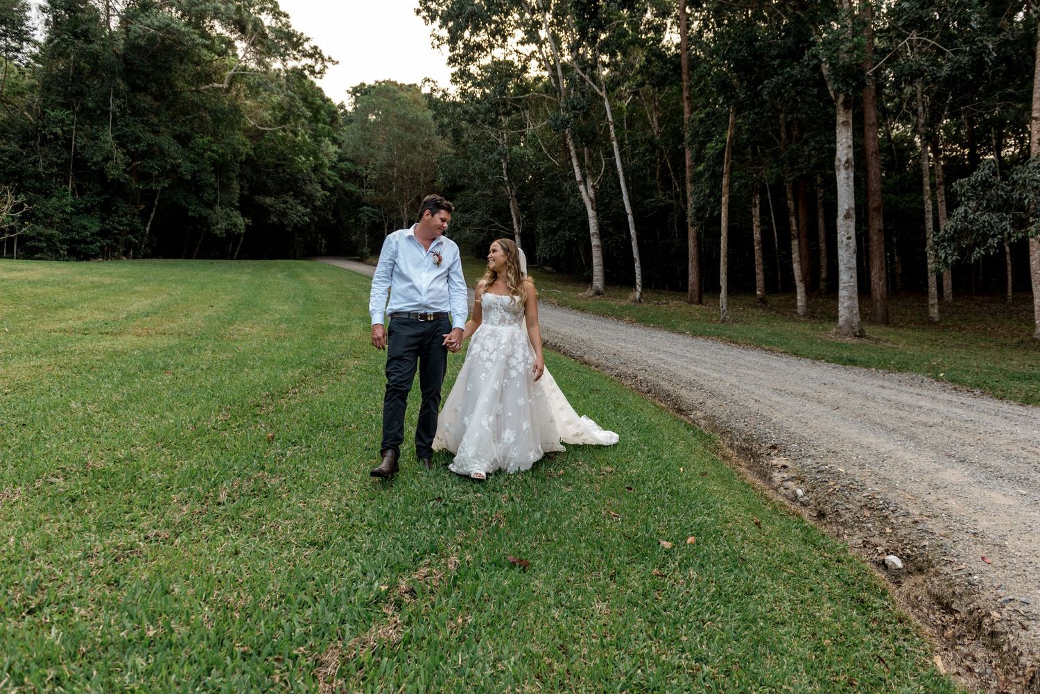 Bride and groom walking hand in hand along a lush green lawn beside a gravel driveway surrounded by tall trees.