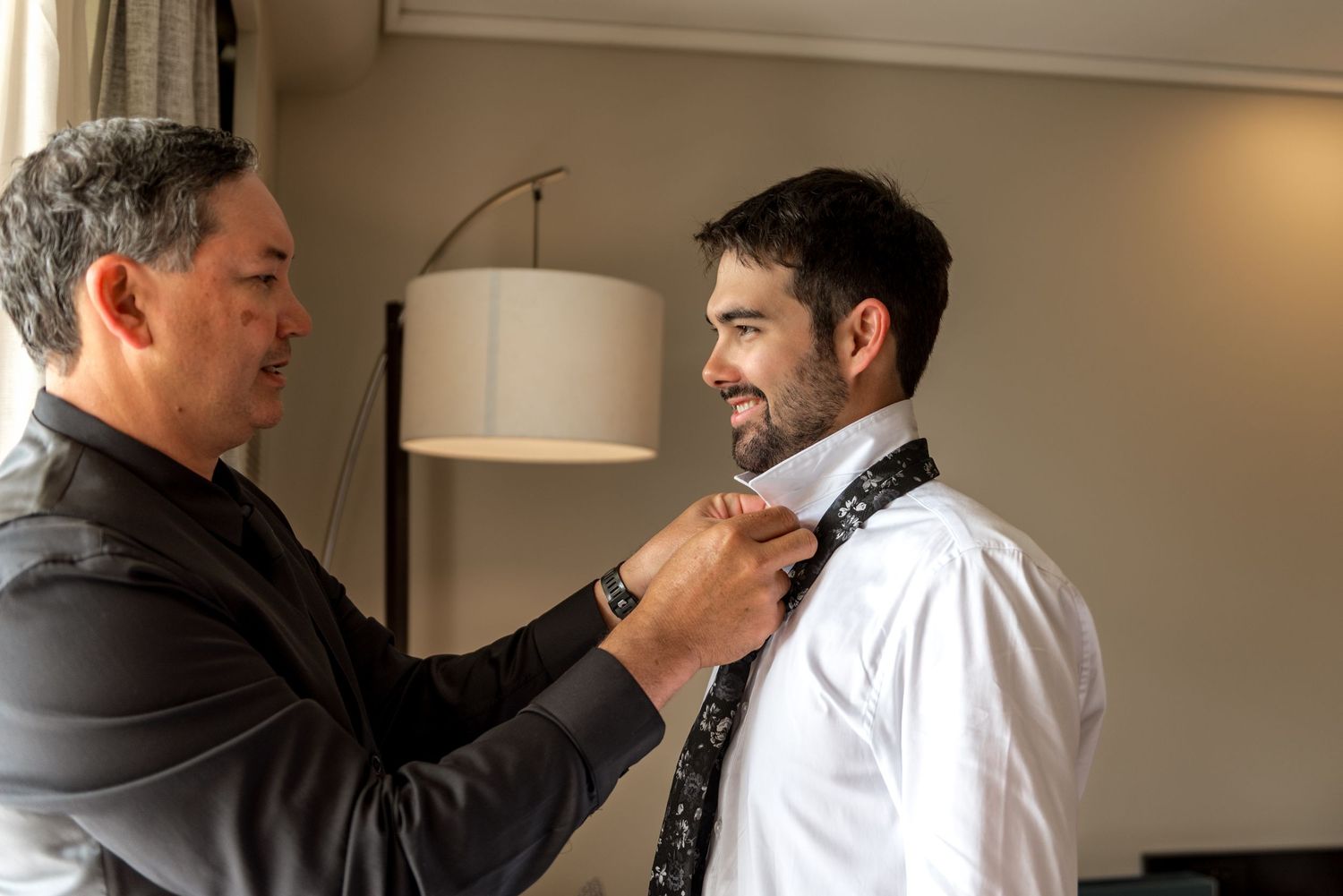 Father helping groom adjust patterned tie while smiling during wedding preparations indoors.