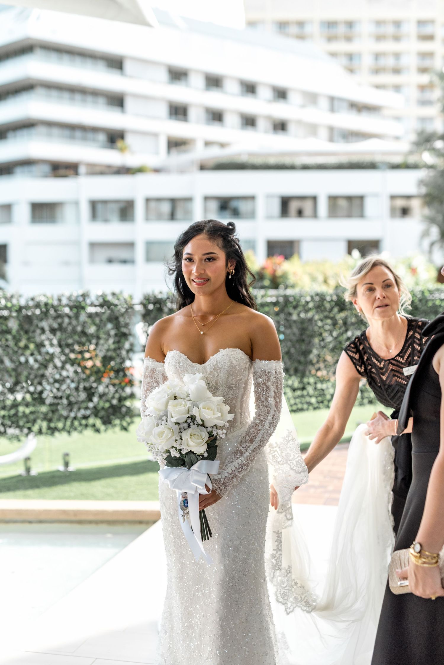 Radiant bride in off-shoulder lace gown carries white rose bouquet at outdoor ceremony.