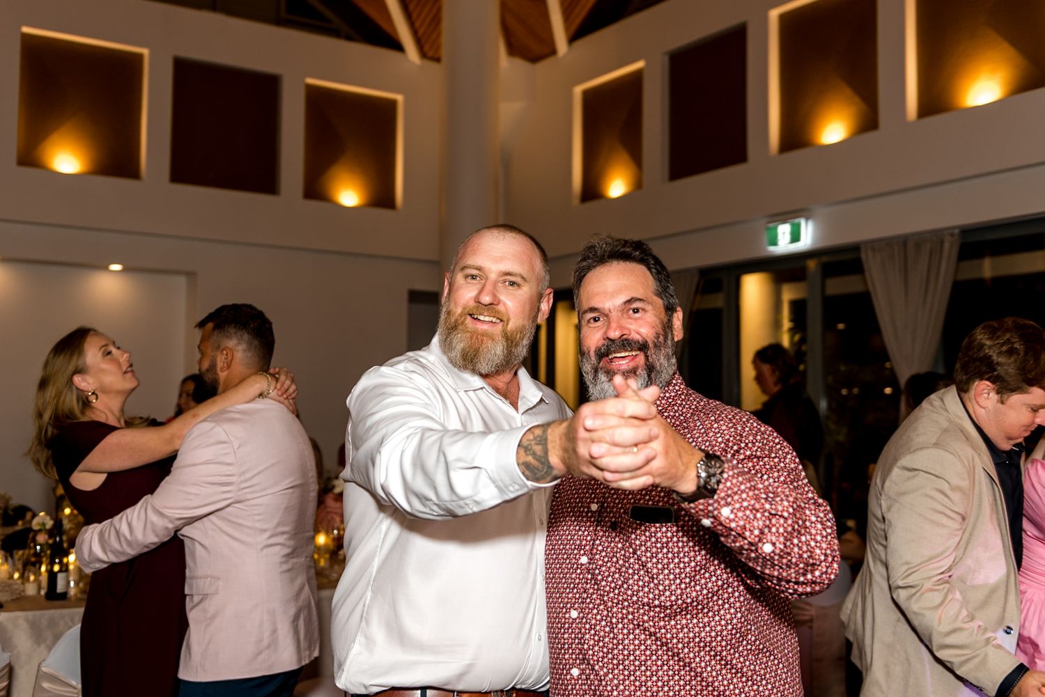 Two bearded men smile and dance together at an elegant wedding reception.