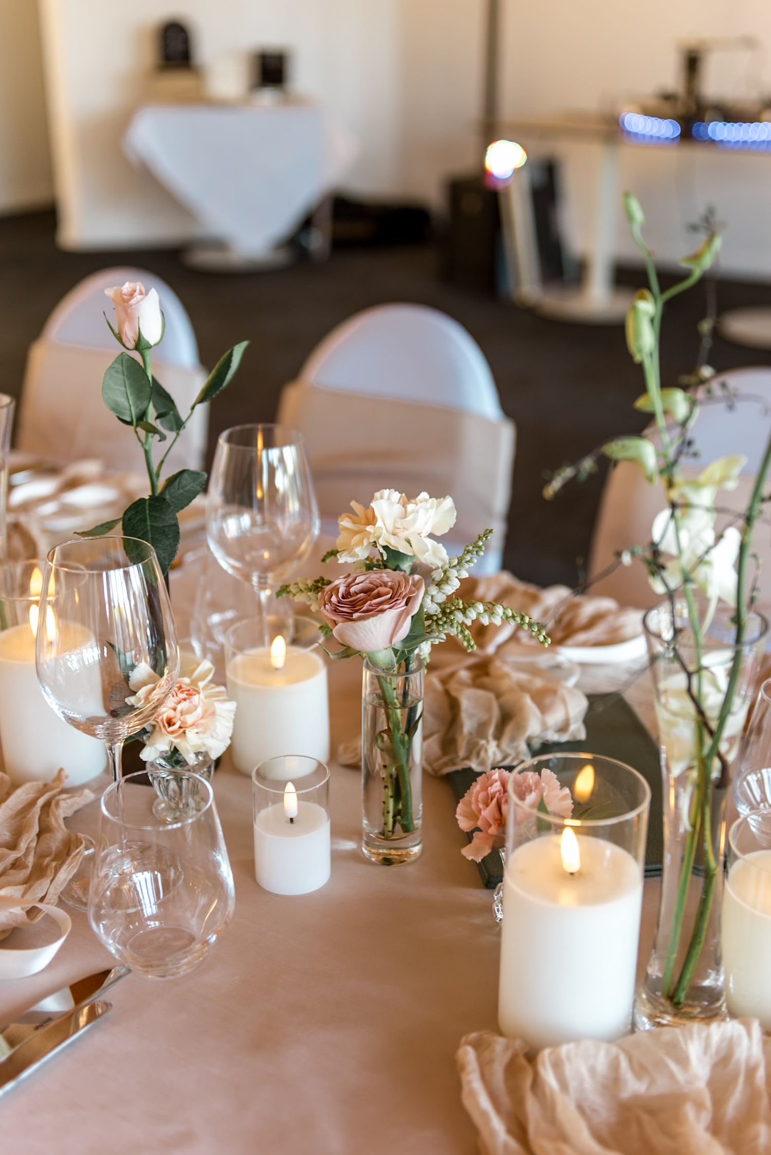 Romantic wedding table with pillar candles, blush roses in bud vases, and crystal glasses on draped linen.